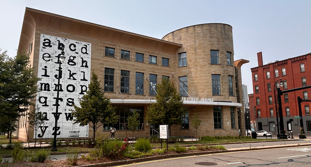 A view of the Worcester Public Library from across the street. There is a large black and white mural of the alphabet in a classic typewriter font that spans all three floors. The rest of the building has large windows, and part of it curves outward to form circular rooms.