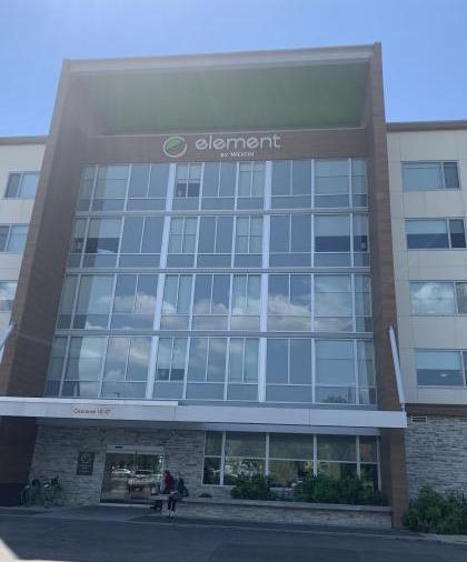 Photo of the front of the five-story, Element hotel. The center tower is framed by an architectural awning and the windows reflect a cloudy sky.