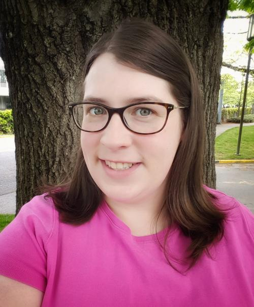 Photo of Crystal Grimes. She is white with brown shoulder-length hair, wearing glasses and a bright pink shirt. She appears to be in a park or similar setting.