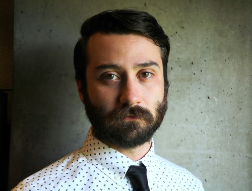 Headshot of J.D. Burns. He has brown eyes and short dark brown hair with a full beard and mustache. He is wearing a white button down shirt with a small black repeating pattern and a black tie and is standing in front of a grey concrete wall.