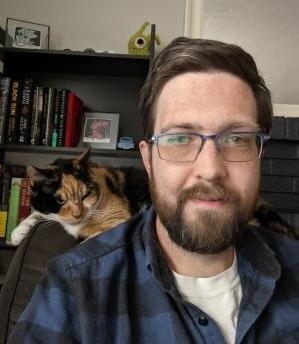 A photo of Sam Lundberg. He is white with brown hair and a short beard. He is sitting in an office with a calico cat on the back of his chair.