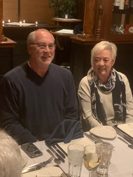 Photo of John and Laura at dinner. John is waring a dark v-neck sweater and smiling at the camera. Laura is wearing a patterned scarf and white sweater, and is smiling at John.