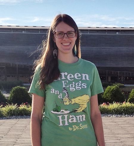 Katharina stands facing the camera. She has long brown hair and is wearing a green Dr. Seuss "Green Eggs & Ham" t-shirt.