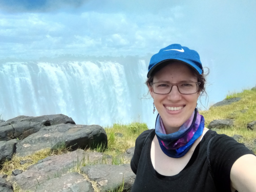 A selfie of Katharina taken in front of a cliff, with a massive waterfall dominating the background. She is smiling and wearing a black t-shirt, blue Nike baseball hat, and a blue and purple neck gaiter.
