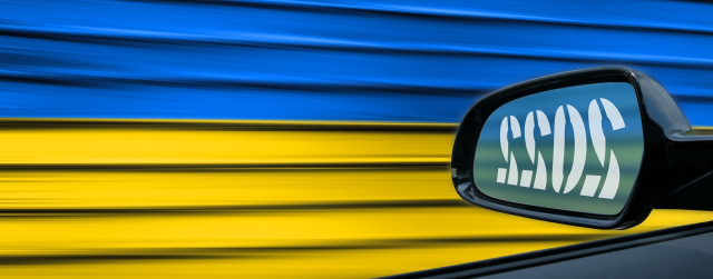 Photo out the driver's window of a car, looking at the side-view mirror. 2022 is written in a large stencil font, in reverse since it's a mirror image. The background is bold blue and yellow with textured stripes as if it is flying past.